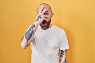 Sarı arka planda dikilmiş İspanyol dövmeli genç bir adam. Şoka girmiş, yüzünü ve gözlerini kapamış, utanmış bir ifadeyle parmaklarından bakıyor.. 