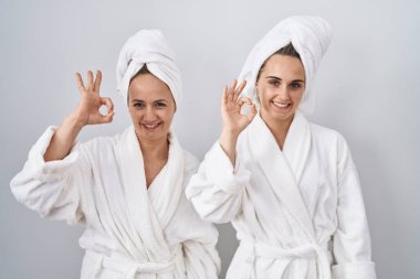 Orta yaşlı kadın ve kızı beyaz bornoz ve havlu giyip gülümsüyor ve el ve parmaklarıyla 