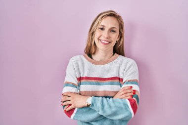Young blonde woman standing over pink background happy face smiling with crossed arms looking at the camera. positive person. 