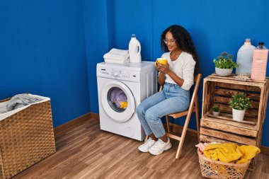 Çamaşır odasında kahve içen genç Latin kadın çamaşır makinesini bekliyor.