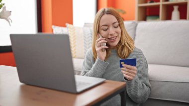 Akıllı telefondan konuşan genç sarışın kadın elinde kredi kartıyla evdeki kanepede oturuyor.