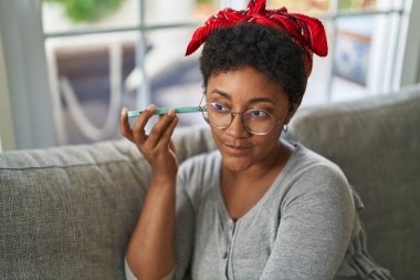 Afro-Amerikalı kadın akıllı telefondan evde kanepede oturmuş sesli mesaj dinliyor.