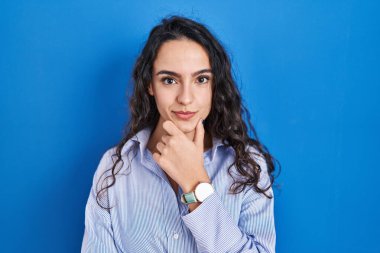 Young brunette woman standing over blue background looking confident at the camera with smile with crossed arms and hand raised on chin. thinking positive. 