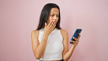 Genç, güzel, İspanyol bir kadın, pembe arka planda sürpriz bir ifadeyle akıllı telefon kullanıyor.