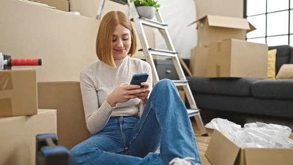 若いですブロンド女性使用してスマートフォン座っている上の床に新しい家 — ストック写真