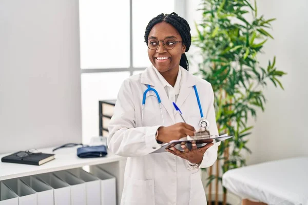 Mujer Afroamericana Doctora Sonriendo Confiada Escribiendo Informe Médico Clínica —  Fotos de Stock