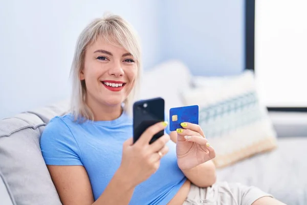 年轻的金发女人坐在家里沙发上 用智能手机和信用卡 — 图库照片