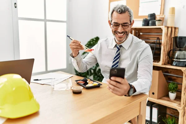 中年灰发男建筑师在办公室用智能手机吃寿司 — 图库照片