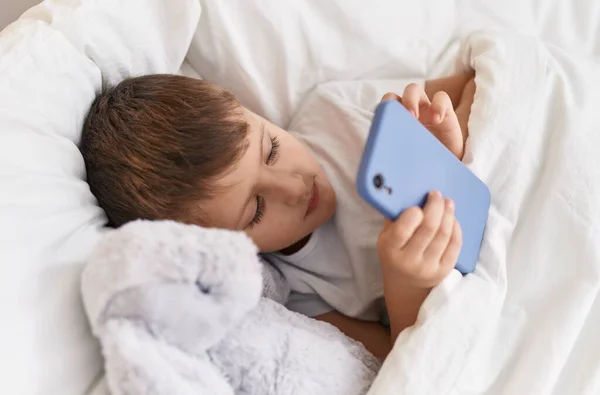 可爱的高加索男孩 用智能手机躺在床上 卧室里挂着洋娃娃 — 图库照片