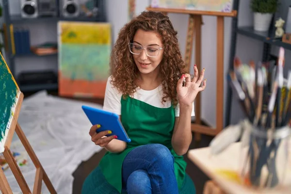 年轻的高加索妇女画家坐在艺术演播室里 用平板电脑做视频通话 用手指签名 微笑友善地摆姿势 展示优秀的象征 — 图库照片