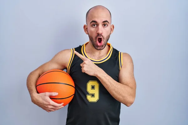 Mladý Plešatý Muž Vousy Basketbalové Uniformě Drží Míč Překvapený Ukazováčkem — Stock fotografie