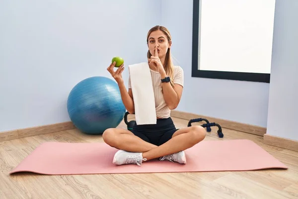 若いブロンドの女性は 唇に指で静かになるように求めて健康的なリンゴを保持スポーツウェアとタオルを身に着けています 沈黙と秘密の概念 — ストック写真