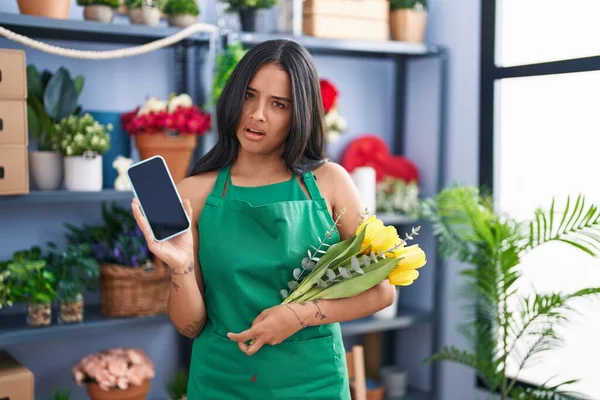 Brunette woman working at florist shop holding smartphone in shock face, looking skeptical and sarcastic, surprised with open mouth