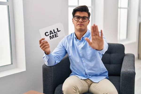 Hispanic man working at therapy office holding call me banner with open hand doing stop sign with serious and confident expression, defense gesture