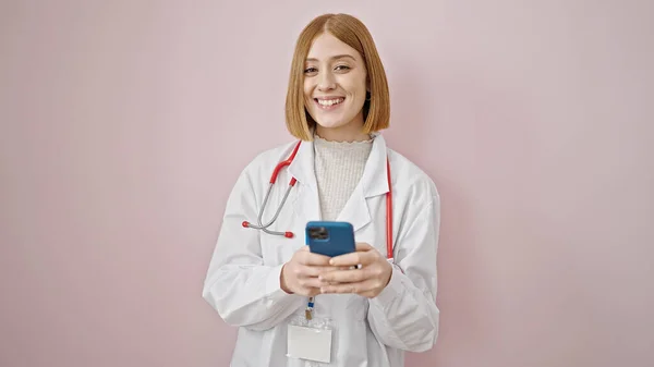 年轻的金发女医生用智能手机在孤立的粉色背景下微笑 — 图库照片