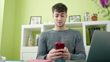 Akıllı telefon ve dizüstü bilgisayar kullanan İspanyol genç yemek odasına üzgün bakıyor.