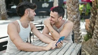 Two men couple smiling confident using smartphone at coffee shop terrace