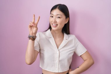 Chinese young woman standing over pink background smiling looking to the camera showing fingers doing victory sign. number two. 