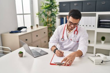 Young hispanic man wearing doctor uniform writing report working at clinic