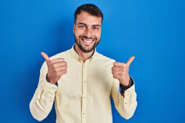 Mavi arka plan başarı işaretinin üzerinde duran yakışıklı İspanyol adam elleriyle olumlu jest yapıyor, gülümsüyor ve mutlu. Neşeli ifade ve kazanan jest. 