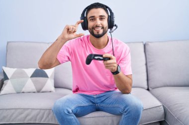 İspanyol asıllı genç adam video oyunu oynuyor kanepeye oturmuş gülümsüyor ve el hareketleriyle küçük boy işareti yapıyor ve kameraya bakıyor. ölçüm kavramı. 