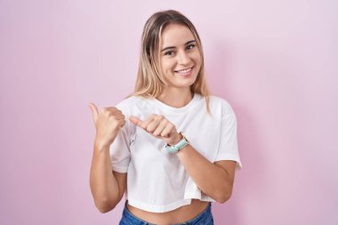 Pembe arka planda duran genç sarışın kadın arka tarafı işaret ediyor. Elleri havada, kendinden emin gülümsüyor. 