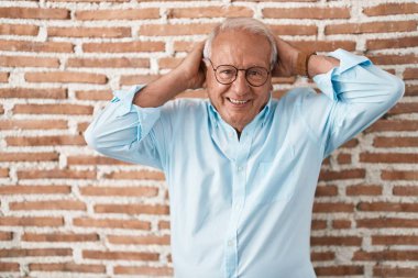 Gri saçlı yaşlı adam tuğlaların üzerinde duruyor rahatlıyor ve esniyor, kolları ve elleri başının arkasında mutlu bir şekilde gülümsüyor. 