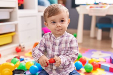 Sevimli beyaz çocuk kreşte yerde kendinden emin bir şekilde otururken gülümsüyor.