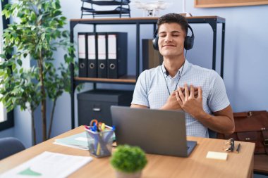 Ofiste çalışan genç İspanyol bir adam kulaklık takıyor elleri göğsünde, gözleri kapalı gülümsüyor ve yüzünde minnet ifadesi var. sağlık kavramı. 