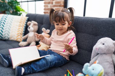 Adorable hispanic girl drawing on notebook sitting on sofa at home