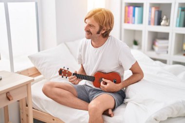 Genç sarışın adam yatakta ukulele oynuyor.