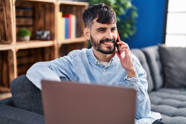 Genç İspanyol adam dizüstü bilgisayar kullanıyor ve evde akıllı telefonla konuşuyor.