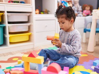 Adorable hispanic girl playing with construction blocks sitting on floor at kindergarten