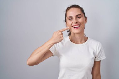 Güzel esmer kadın izole edilmiş arka planda duruyor. Parmaklarıyla yüzünü ve burnunu gösteriyor. Neşeli bir şekilde gülümsüyor. güzellik konsepti 