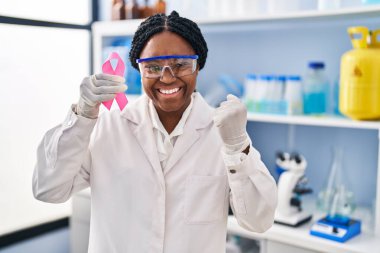 Afrika kökenli Amerikalı bir kadın, elinde pembe kurdeleyle laboratuvarda çalışıyor. Gururla bağırıyor, zaferi kutluyor ve kollarını kaldırarak başarıyı kutluyor. 
