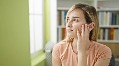 Genç sarışın kız öğrenci kütüphanede masada oturmuş düşünüyor.