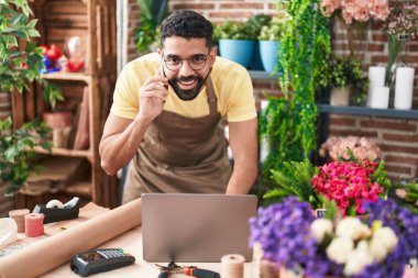 Young arab man florist talking on smartphone using laptop at florist