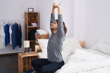 Young hispanic man waking up stretching arms at bedroom