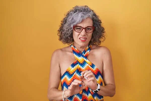 Middle age woman with grey hair standing over yellow background disgusted expression, displeased and fearful doing disgust face because aversion reaction.