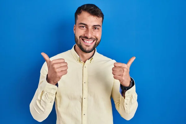 青い背景の成功の印の上に立つハンサムなヒスパニック系の男性は手で積極的なジェスチャーを行うと 親指を笑顔と幸せ 陽気な表情と勝者のジェスチャー — ストック写真