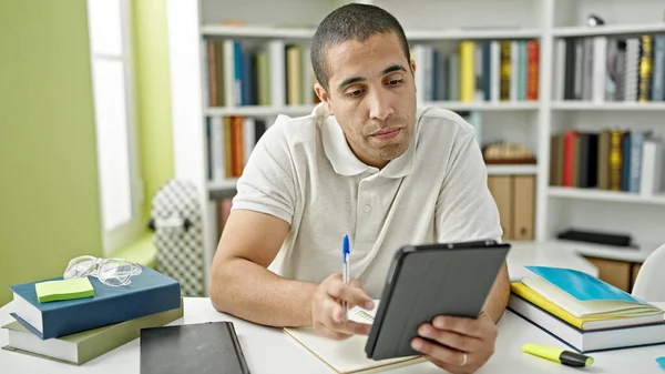 Young hispanic man student using touchpad studying at library university