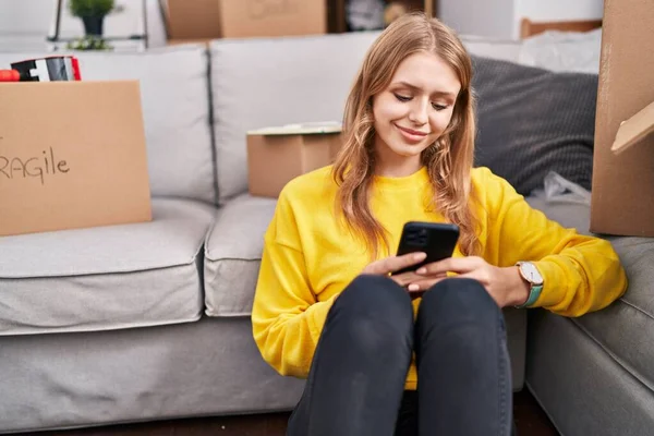 年轻的金发女人用智能手机坐在新家的地板上 — 图库照片