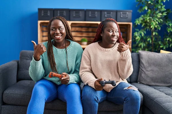两个非洲女人在玩电子游戏 手里拿着控制器 把大拇指指向一边 开心地张开嘴 — 图库照片