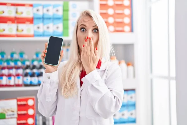 在药店工作的白人妇女手拿着智能手机屏幕捂住嘴 感到震惊和害怕犯错 惊讶的表情 — 图库照片