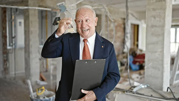 stock image Senior grey-haired man real state agent holding clipboard and keys of new house at construction site