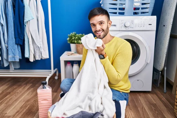 年轻的惊慌失措的男人在洗衣房自信地笑着闻到干净衣服的味道 — 图库照片