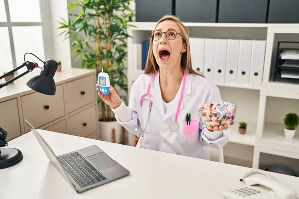 Young blonde doctor woman holding glucometer and sweets angry and mad screaming frustrated and furious, shouting with anger looking up.