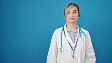 Genç sarışın kadın doktor gülümsüyor kendine güvenen, izole edilmiş mavi arka planda sunan.
