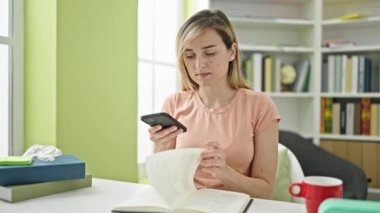 Kütüphanede akıllı telefon okuma kitabı kullanan genç sarışın kız öğrenci.