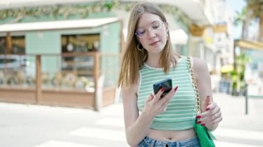 Genç, sarışın, kendine güvenen bir kadın kahve dükkanının terasında akıllı telefondan konuşuyor.